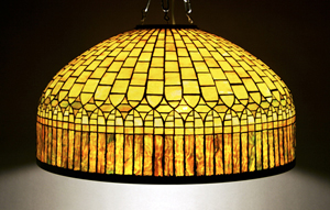 Tiffany Studios Favrile glass and bronze curtain-border lamp shade, 1899-1920, the broad domical shade composed of geometric glass tiles in heavily mottled pale olive-green and opalescent glass, above a lower border of vertical rectangular sections in striated crimson and golden-amber opalescent ice-textured glass; shade impressed TIFFANY STUDIOS NEW YORK 1575. Diameter of interior 24 7/8 inches. Estimate $30,000 to $50,000. Image courtesy Leslie Hindman Auctioneers.