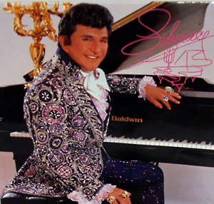 In this photograph, Liberace is dressed to entertain in a dazzling stage costume, his fingers bedecked with gold and diamond jewelry. Mid-Hudson Galleries image.