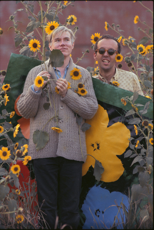 Andy Warhol in a field of black-eyed Susans with Taylor Mead holding an early “Flowers” canvas as a backdrop in Queens, New York. Title: Warhol Flowers VII Location: Flushing, Queens Medium: Chromogenic Print Edition: 60 with 7 Artist Proofs Size: 24 x 16 inches Executed: 1964  Printed: 2012 Copyright 2010 William John Kennedy / Courtesy of Kiwi Arts Group
