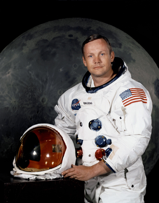 Portrait of Astronaut Neil A. Armstrong (American, 1930-2012), commander of the Apollo 11 Lunar Landing Mission in his space suit, with his helmet on the table in front of him. Behind him is a photograph of the lunar surface. NASA Photo  No. S69-31741, taken July 1, 1969.