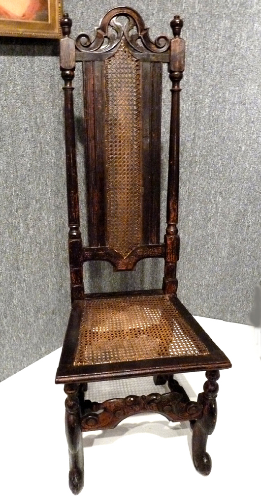 Close by is a close relative of the William and Mary chair, a Flemish-style chair from New England, first quarter of the 18th century, from the Dows. This chair, while showing the long lean look of the William and Mary style, retains the carved crest and stretcher of the earlier Restoration style of 1660 when the monarchy was restored in England. Very few examples of this type chair were made in the Colonies. Most were imported from London.