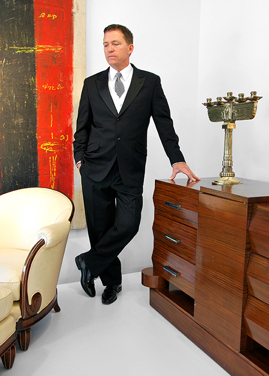 Palm Beach Modern’s auctioneer, Rico Baca, gets into the Gatsby mood as he inspects premier items in the company’s May 25 auction, including (clockwise from left) one of a pair of Jules Leleu French Art Deco period chairs with ottomans, a large Jeff Compertz abstract painting, one of a pair of circa-1930 French Art Deco Egyptian Revival candelabra, and an Alfred Porteneuve French Art Deco rosewood with silver-plated bronze sideboard/dresser made in 1939 for the SS Pasteur ocean liner. Palm Beach Modern Auctions image.