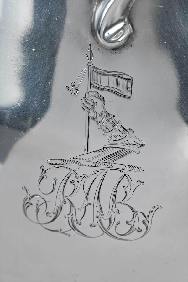 Detail from a large George III English silver pitcher by Hester Bateman, showing her engraving of a heraldic design with an arm holding a flag above the initials RAB. The pear-form pitcher made $3,500 plus the buyer’s premium in May 2022. Image courtesy of Brunk Auctions and LiveAuctioneers.